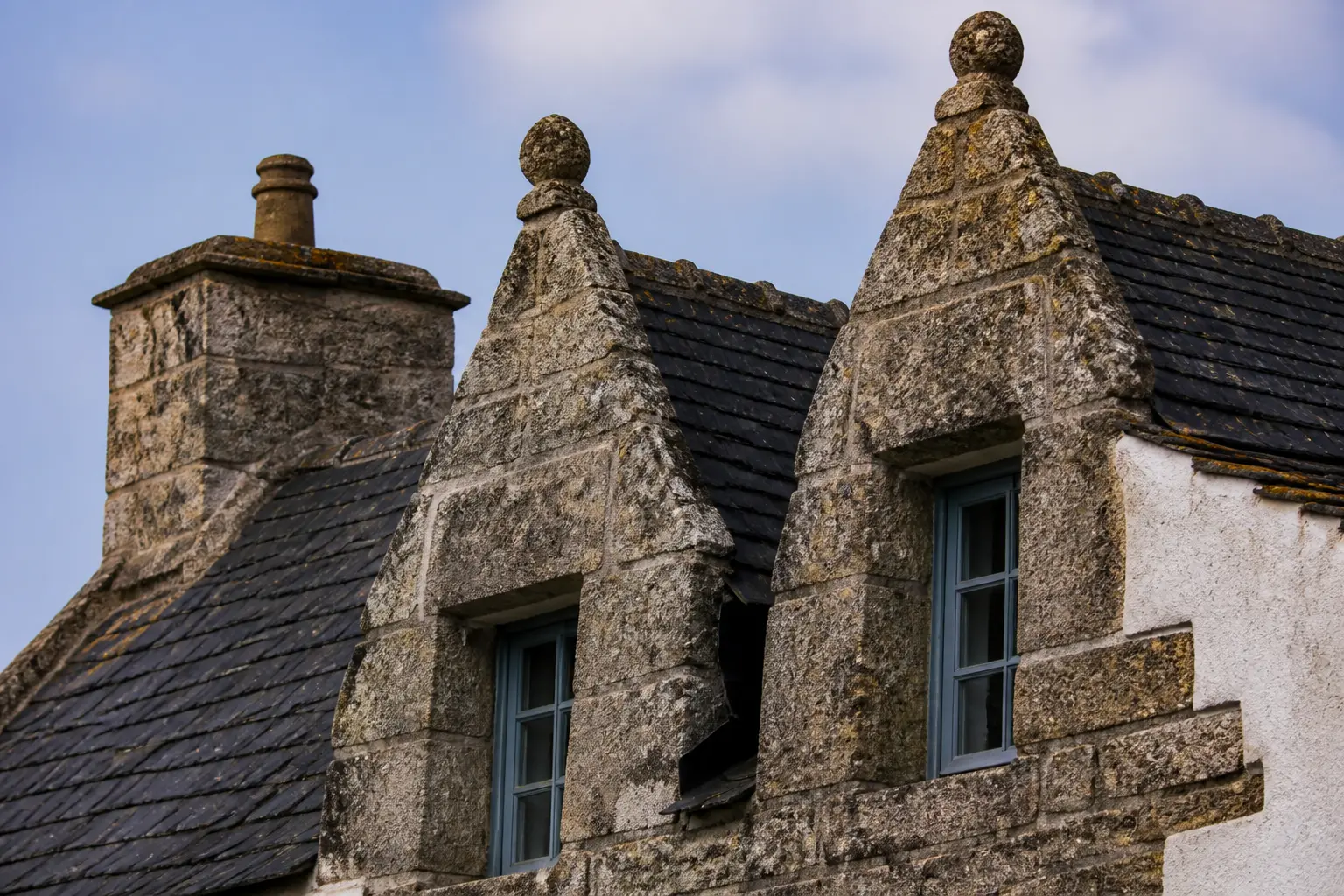 Maison bretonne en granit gris, toit d’ardoise pentu, fenêtres à encadrements de pierre et cheminée.